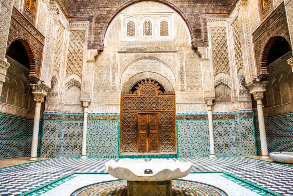 Porta in cedro scolpito della sala di preghiera della madrasa Al Attarine, incorniciata da colonne di marmo e zellige, con la vasca in primo piano