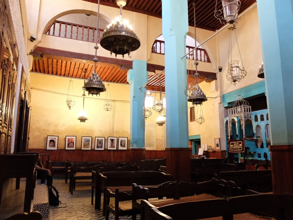Innenraum der Synagoge Ibn Danan in Fès mit türkisfarbenen Pfeilern, hölzerner Empore, hängenden Leuchtern und Bänken